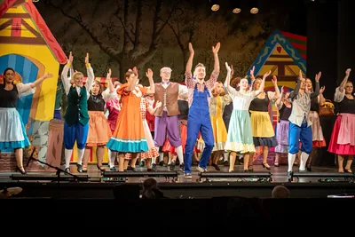 A colourful pantomime ensemble mid-routine with arms raised on a painted fairytale village set